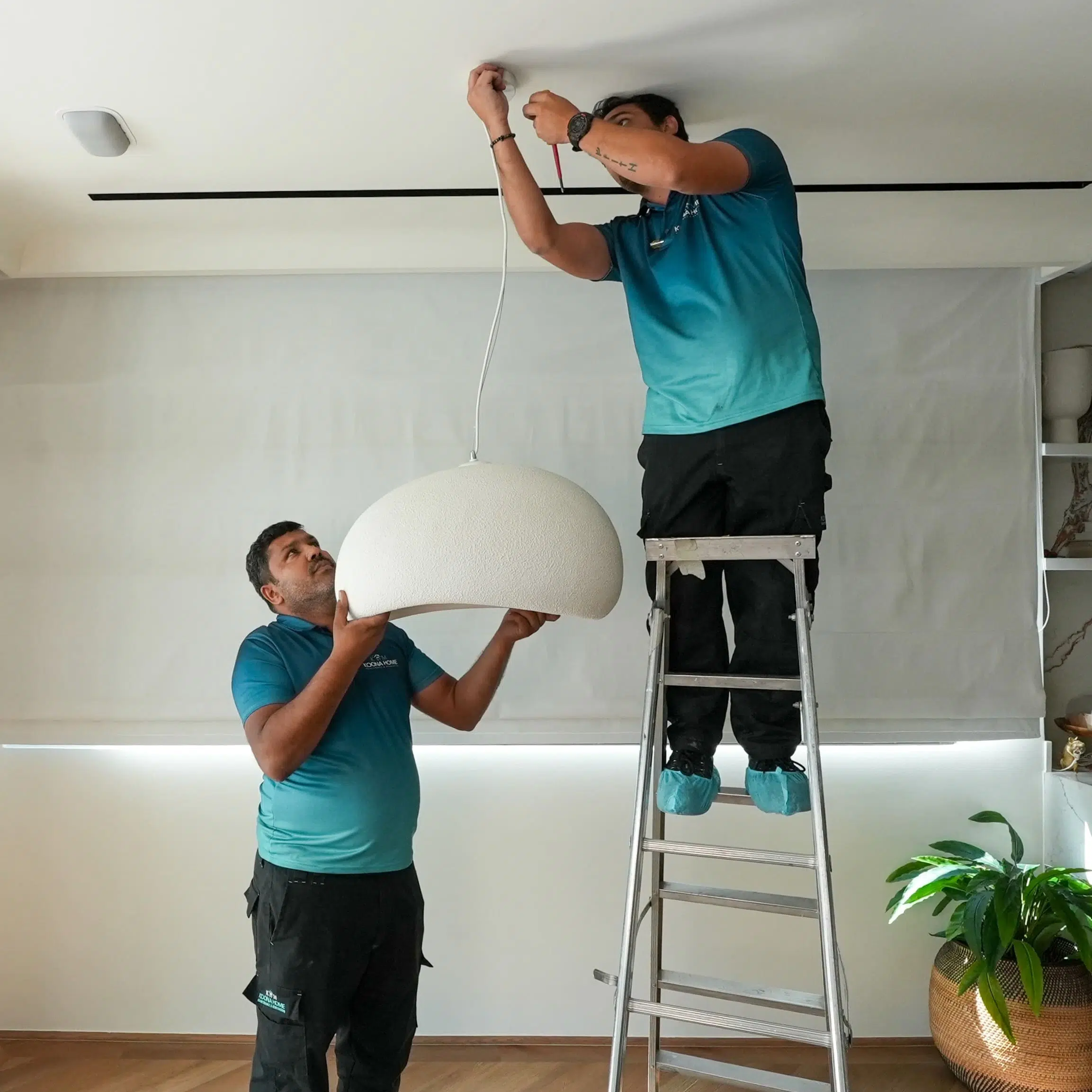 Move in and out handyman services in Dubai technicians installing a ceiling light fixture, one standing on a ladder connecting wires while the other supports the lamp in a modern home interior.